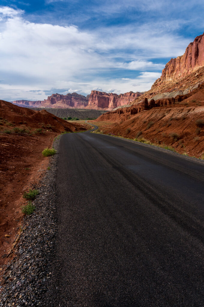 Winding road through rugged red rock canyon with towering cliffs and partly cloudy sky background Wallpaper