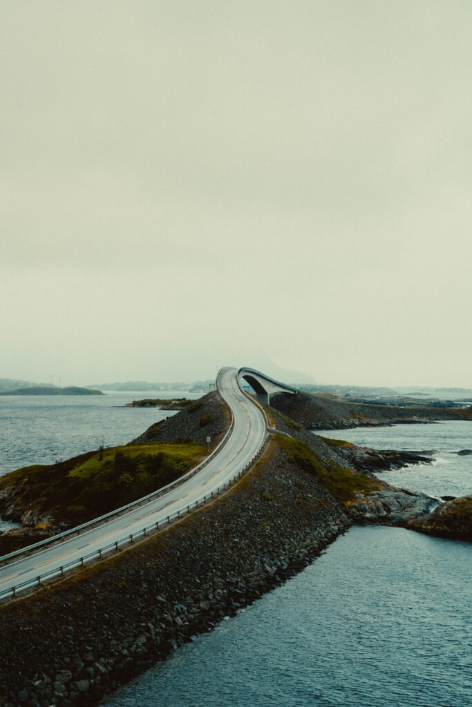 Winding road and arched bridge over blue water with lush landscape under overcast sky wallpaper