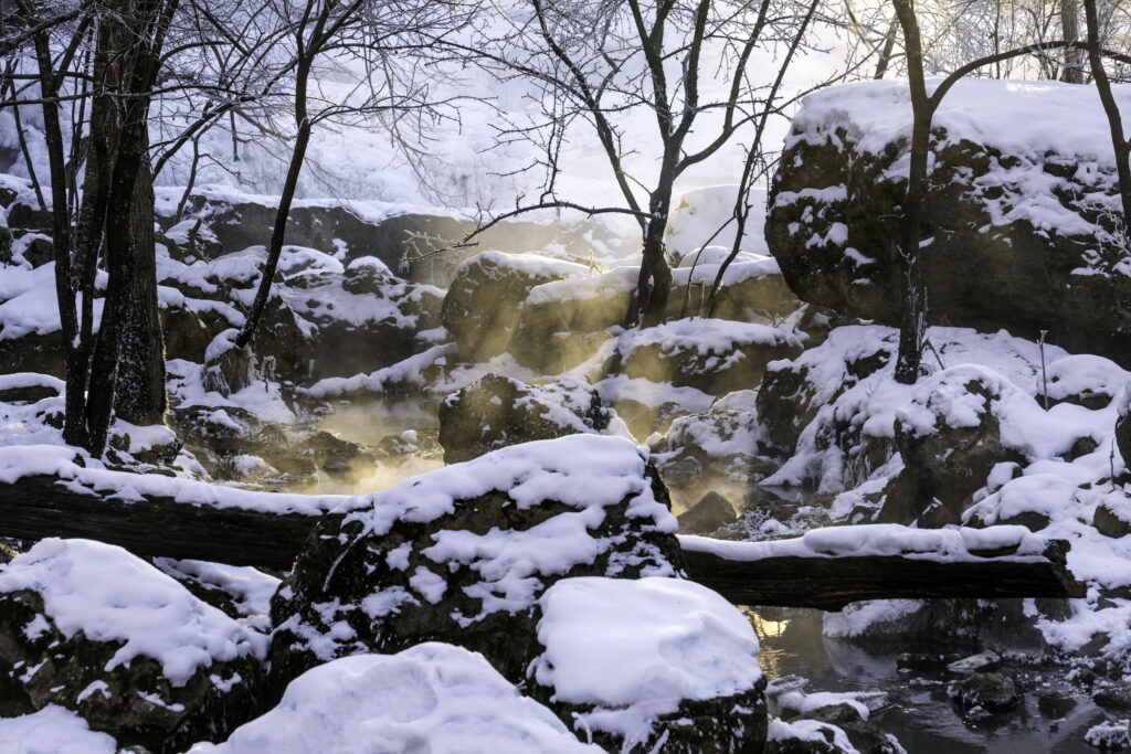 Sunlight shining through bare trees on snowy rocks with steam rising from a winter creek Wallpaper