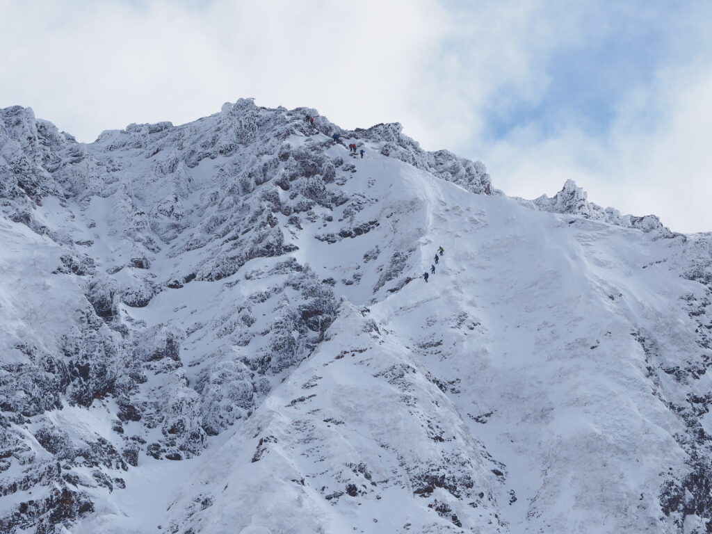 Group hiking up rugged snow-covered mountain slope under partly cloudy sky winter wallpaper background