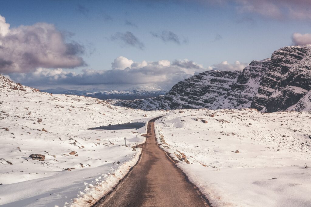 Winding road through snow-covered landscape toward rugged mountains under cloudy sky wallpaper