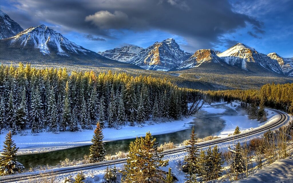 Winter landscape wallpaper with snowy railway by river and fir trees under dramatic mountains