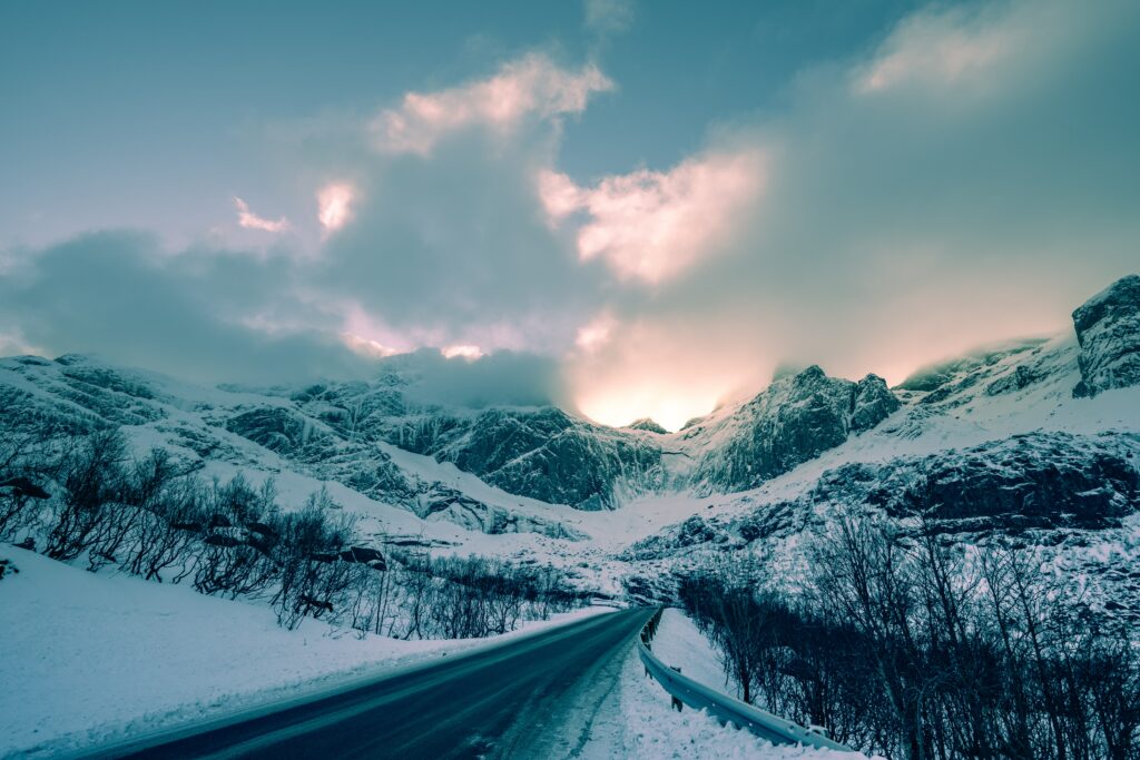 Winter road winding through snow-covered Norwegian mountains under dramatic clouds and sunlight Wallpaper