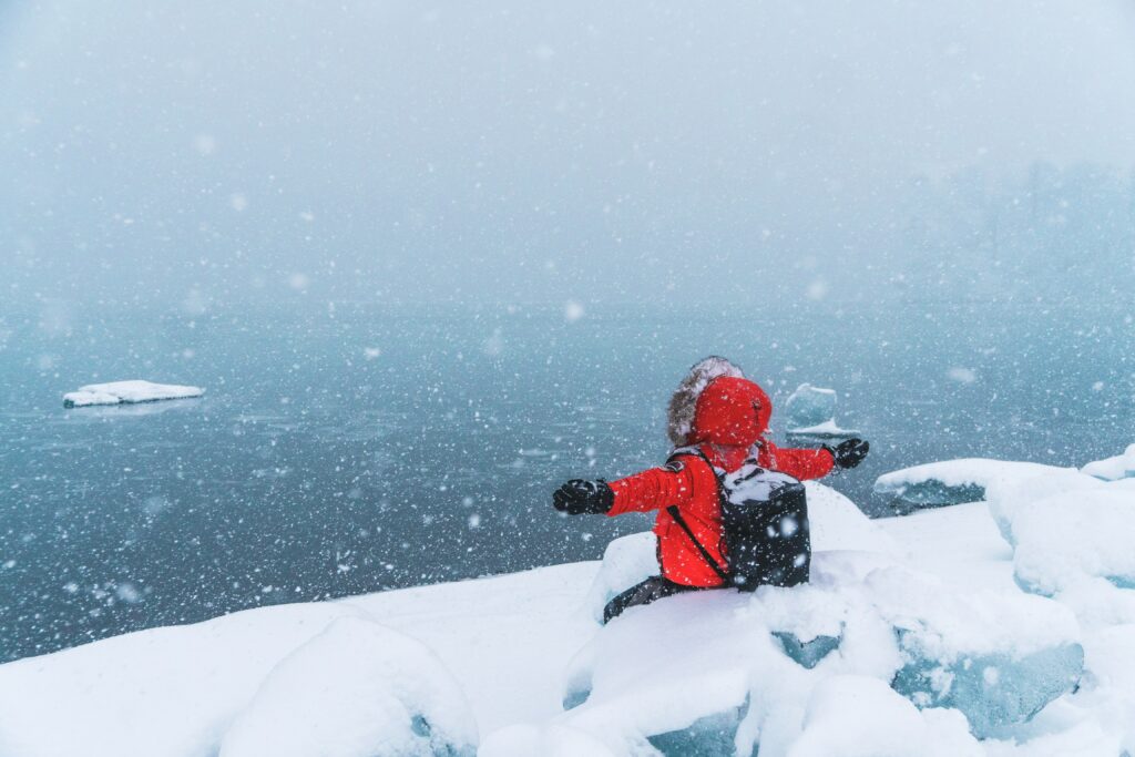 Person in red jacket sits in fresh snow by lake arms outstretched snowflakes falling winter scene Wallpaper