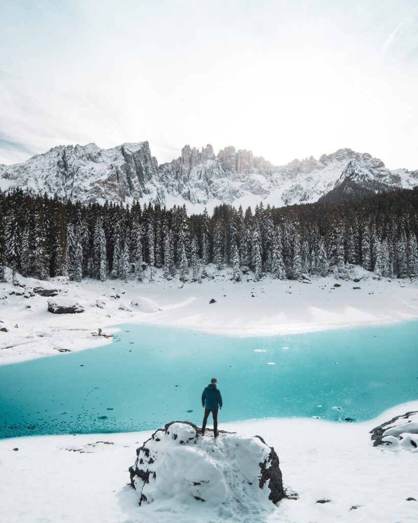 Lone man on snowy rock gazing at turquoise lake pine trees and snow-covered mountains winter scene Wallpaper