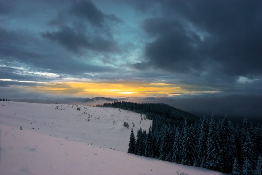 Winter wallpaper with snow-covered mountains pine forest and warm sunset glow under dramatic sky