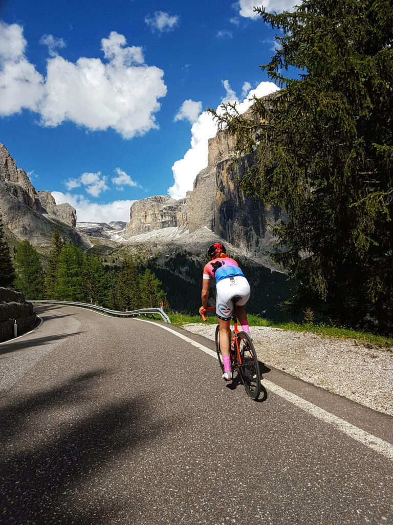 Woman cycling on winding Alpine road with mountains pine trees and bright blue sky background Wallpaper