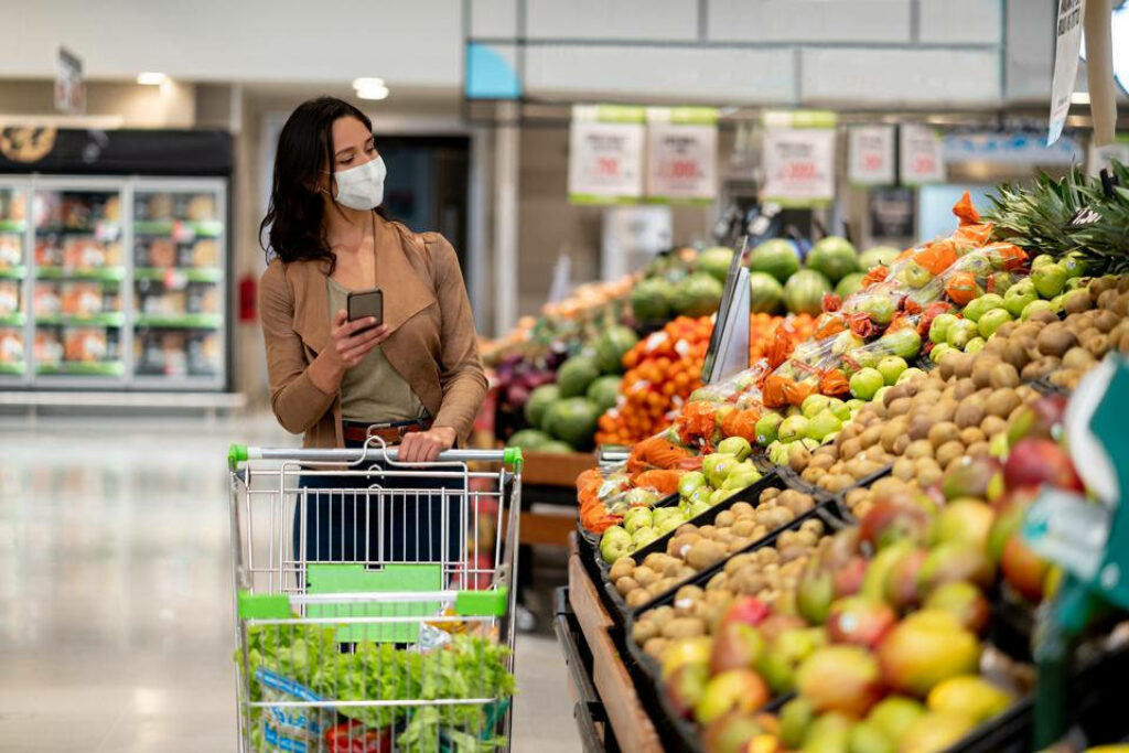 Masked Woman Grocery Shopping: Fresh Produce & Smartphone Use Wallpaper