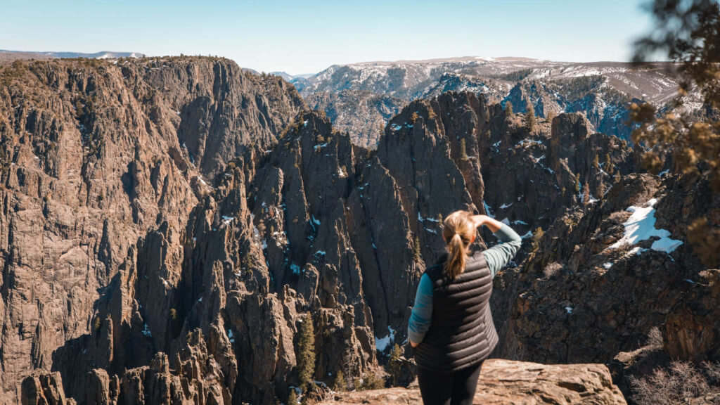 Woman on rocky cliff overlooking Black Canyon rugged spires steep walls snow patches clear sky Wallpaper