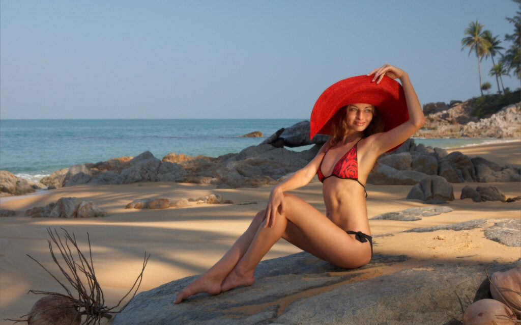 Woman in red hat and bikini posing on sunlit beach with calm sea and rocky shore background Wallpaper