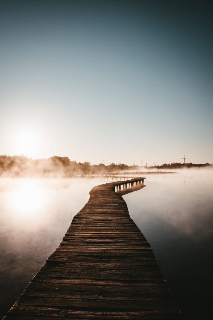 Wooden bridge over calm water in soft morning fog with warm sunlight creating serene mood Wallpaper