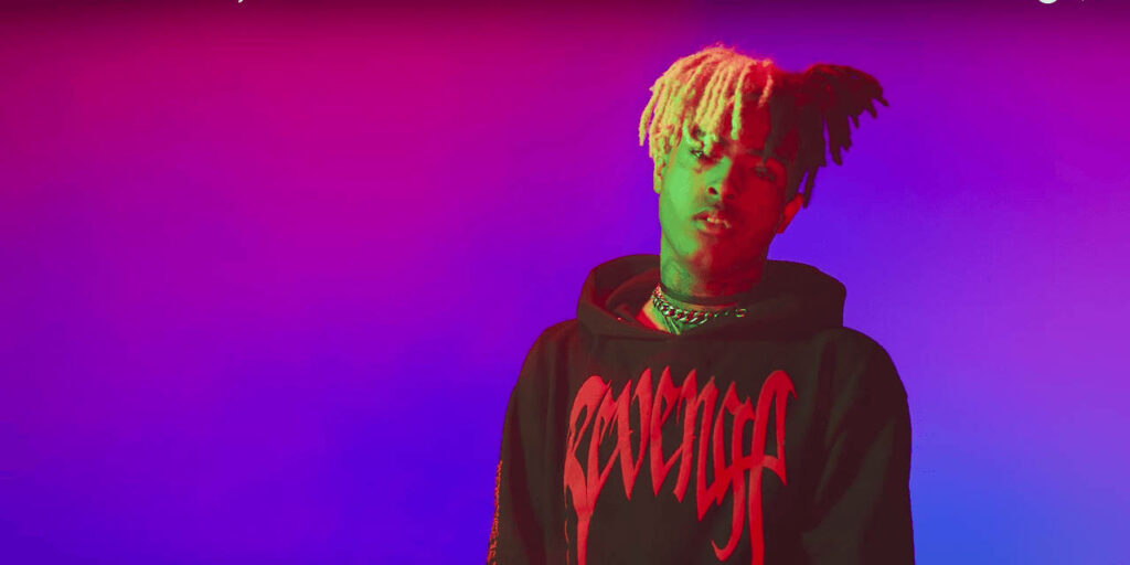 Portrait of rapper XXXTentacion in black Revenge hoodie with blonde hair on purple blue gradient background Wallpaper