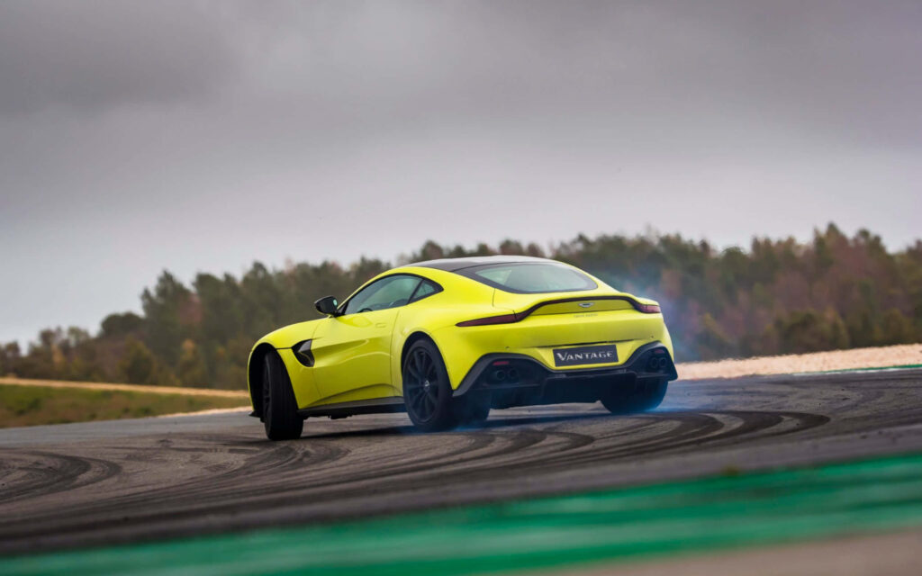 Yellow Aston Martin Vantage drifting on racetrack with cloudy sky and green landscape background Wallpaper