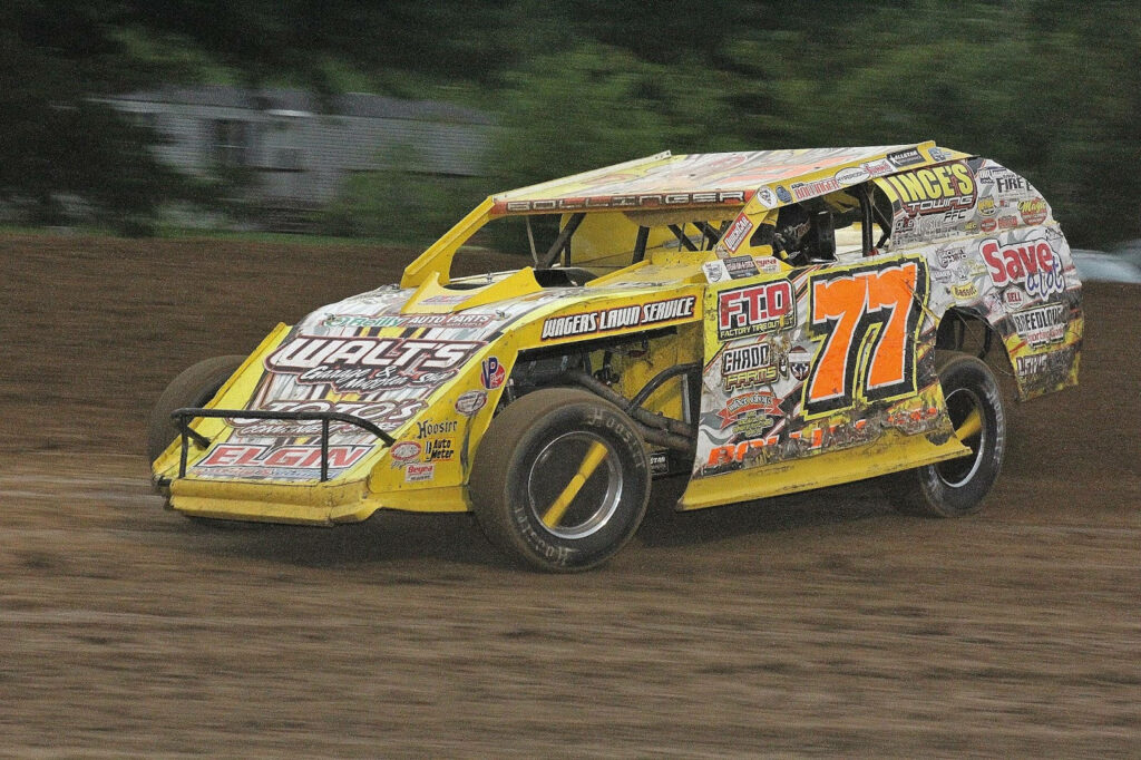 Yellow dirt car number 77 with sponsor decals racing fast on muddy dirt track action wallpaper