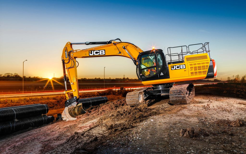 Yellow JCB JS300c excavator on dirt mound near drainage pipes with sunset and traffic light trails Wallpaper