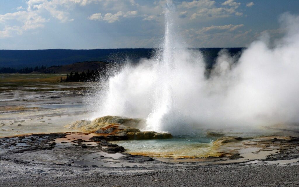 Yellowstone geyser erupting steam and water over mineral-rich ground with rolling hills background Wallpaper