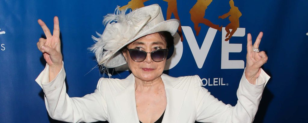 Yoko Ono peace sign pose with white hat dark sunglasses blue Cirque du Soleil LOVE backdrop wallpaper