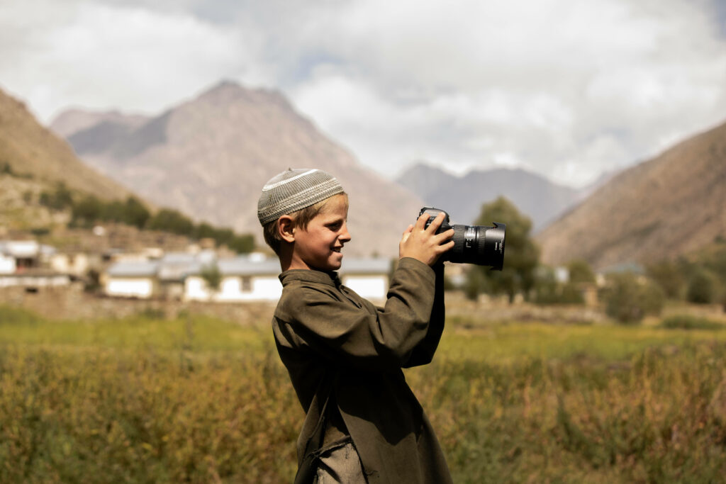 Young boy in traditional Afghan clothes happily photographing scenic mountain village landscape Wallpaper