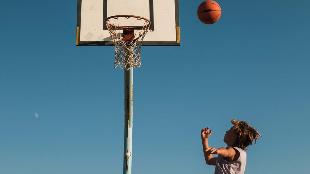 Young basketball player mid-air shooting at outdoor hoop with clear blue sky and crescent moon background Wallpaper