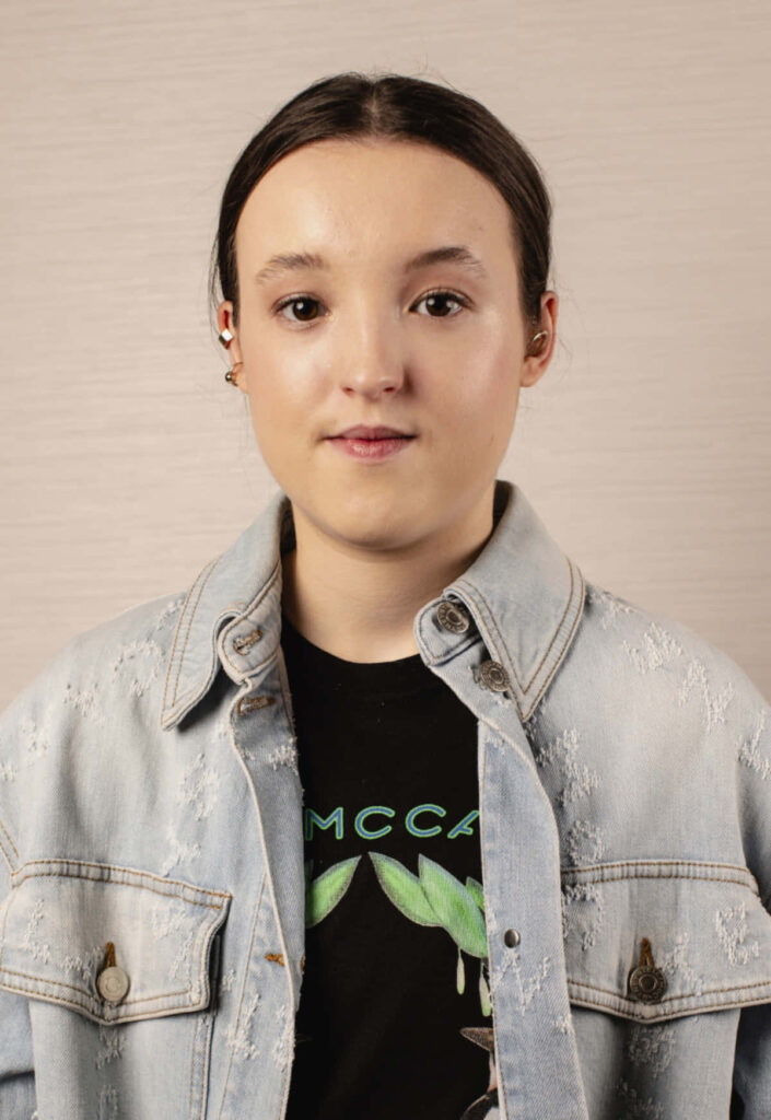 Young Bella Ramsey in light denim jacket and black graphic tee on neutral modern wallpaper background