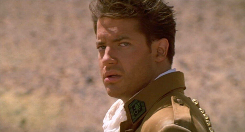 Young Brendan Fraser in rugged tan military jacket against dramatic desert for action film wallpaper