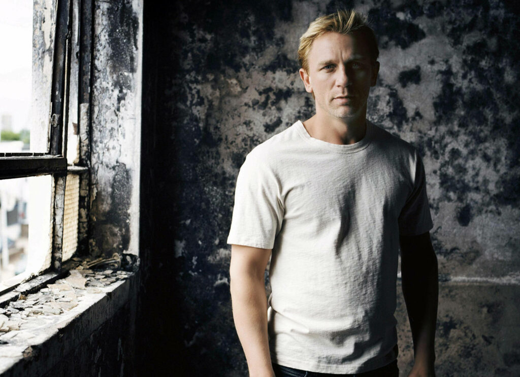 Young Daniel Craig in white tee by weathered window natural light highlights rugged wall portrait Wallpaper