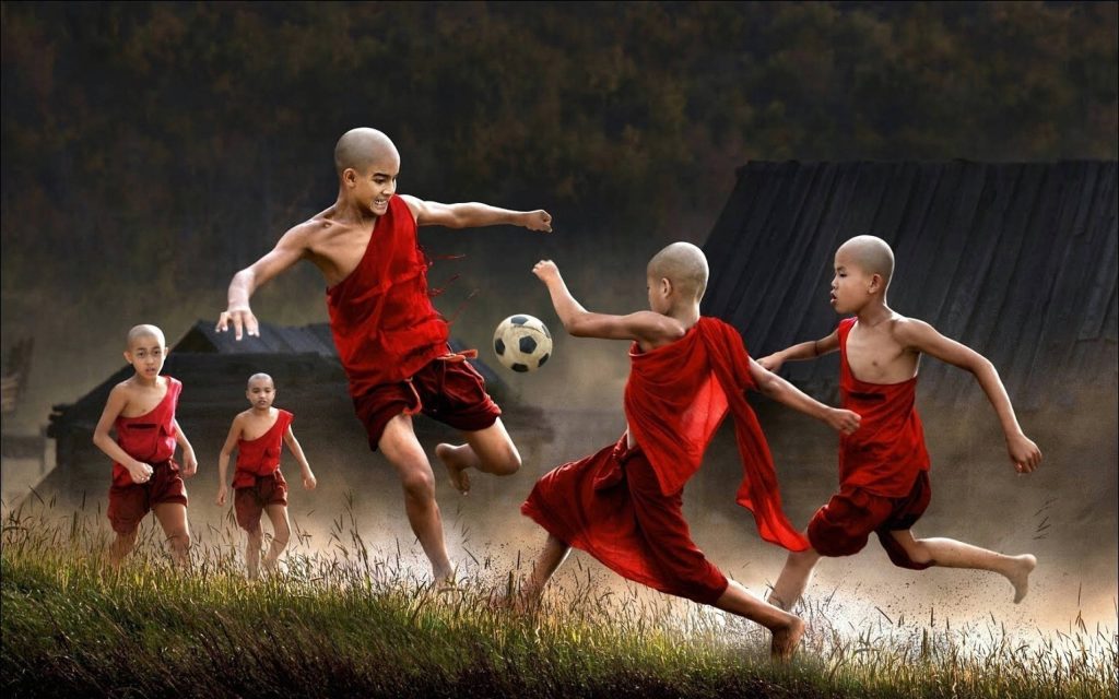 Young monks in bright red robes play soccer on rustic field vibrant dynamic football match wallpaper
