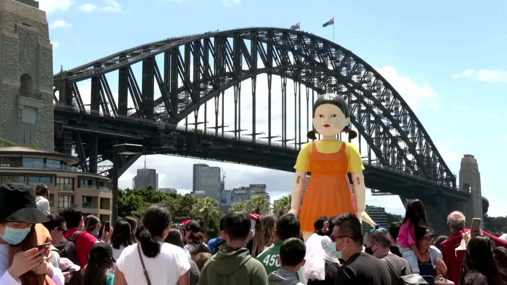 Spectacular Sydney Scene: Young-hee Doll Statue amidst a Sea of Participants at Squid Game Red Light, Green Light Event near Sydney Harbour Bridge Wallpaper