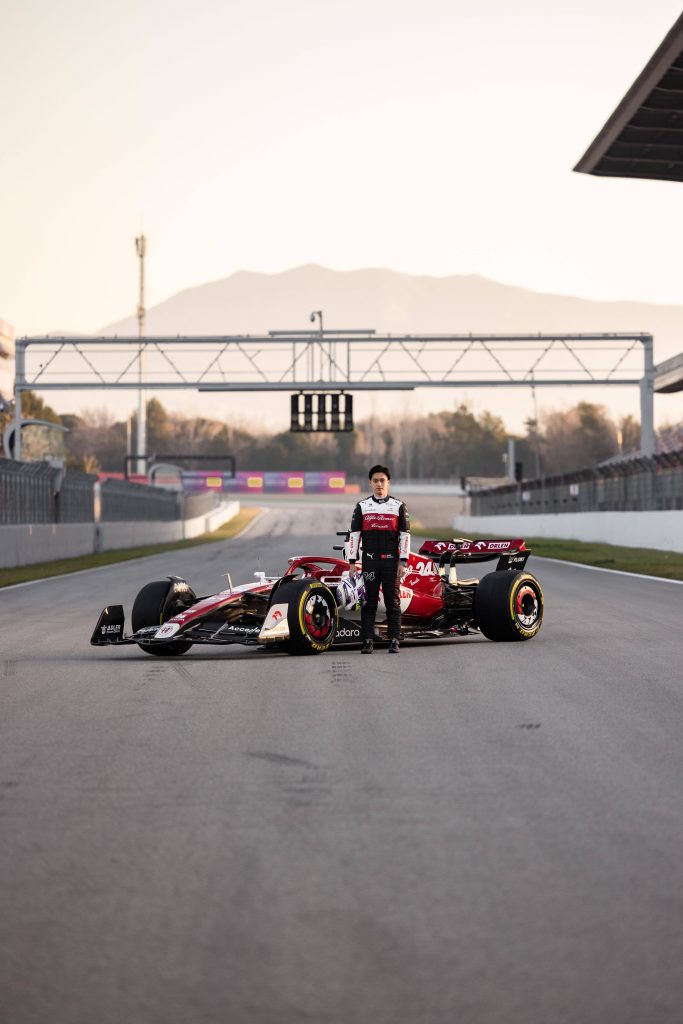 F1 driver Zhou Guanyu portrait beside Alfa Romeo car at sunset on empty track cinematic wallpaper background