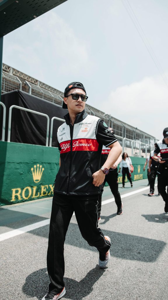 F1 driver Zhou Guanyu in Alfa Romeo gear and sunglasses walking past Rolex wall in sunny paddock Wallpaper