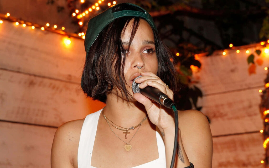 Zoe Kravitz performing live wearing layered necklaces and green cap with vibrant string lights background Wallpaper